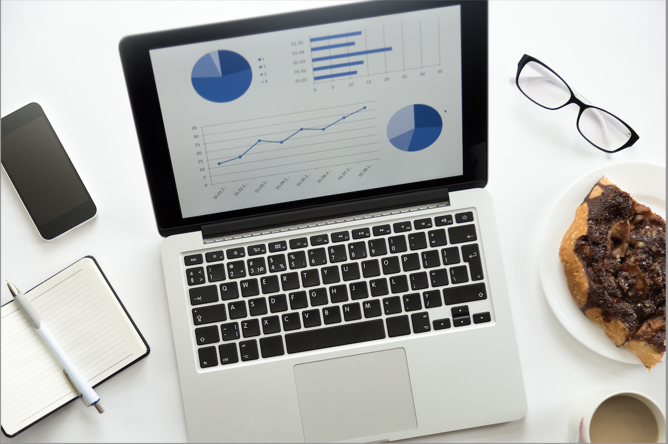 Image: A modern, clean workspace featuring a laptop with data analytics on the screen. Graphs and pie charts present business performance metrics. Nearby, a smartphone, eyeglasses, a cup of coffee with a half-consumed cup